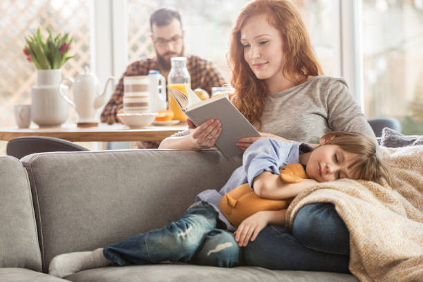 Mãe lendo no sofá com o filho dormindo no colo, e o pai à mesa ao fundo